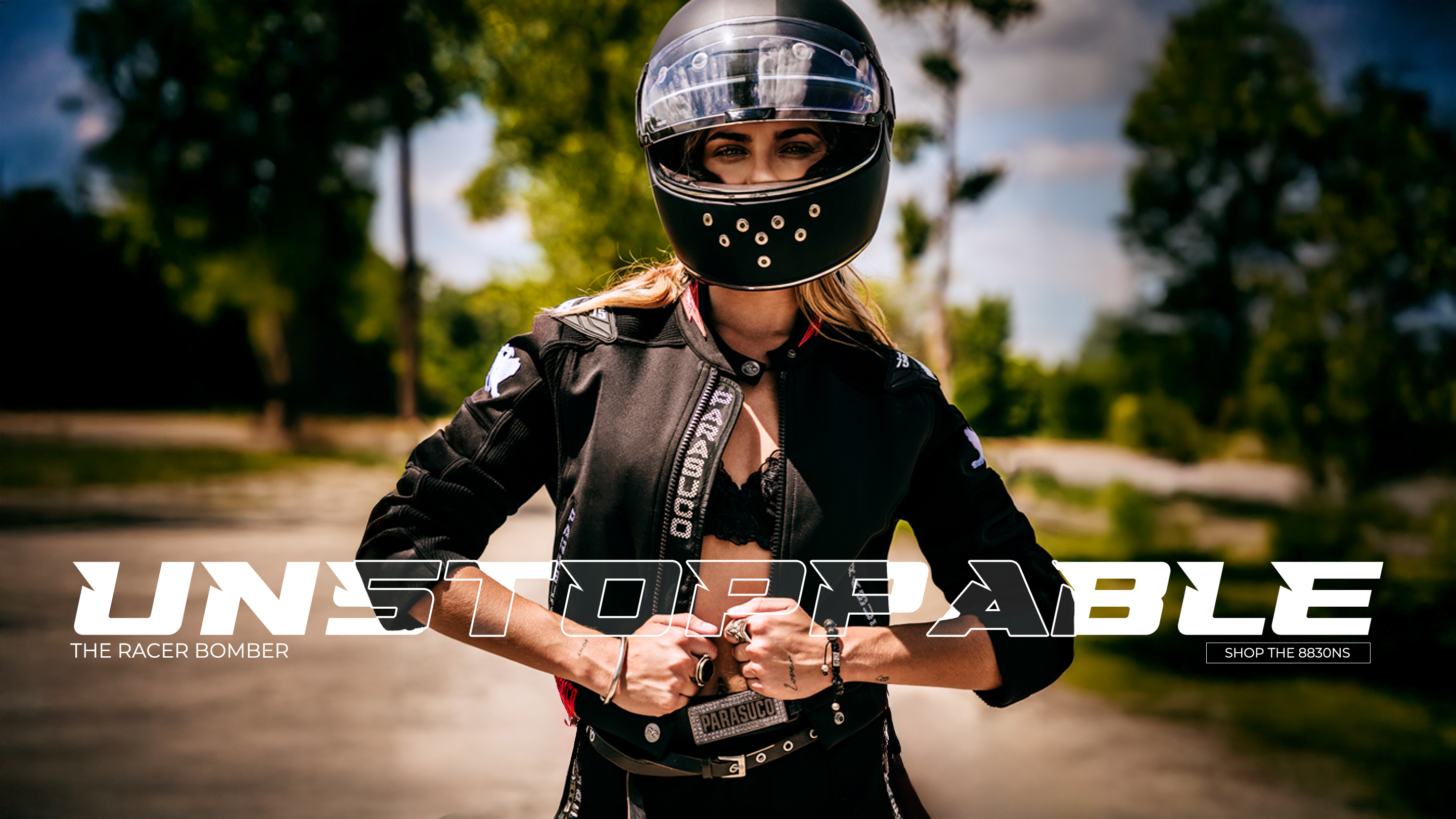 
A muscular man with short hair and tattoos stands in profile, leaning on a rustic wooden fence. He is wearing a black, sleeveless top with a white graphic print, black leather pants, and red motorcycle boots. He holds a red and black helmet in his left hand. The background is a dirt road bordered by trees, with sunlight filtering through the leaves. The image has a dark, vertical overlay on the left side with the text 
