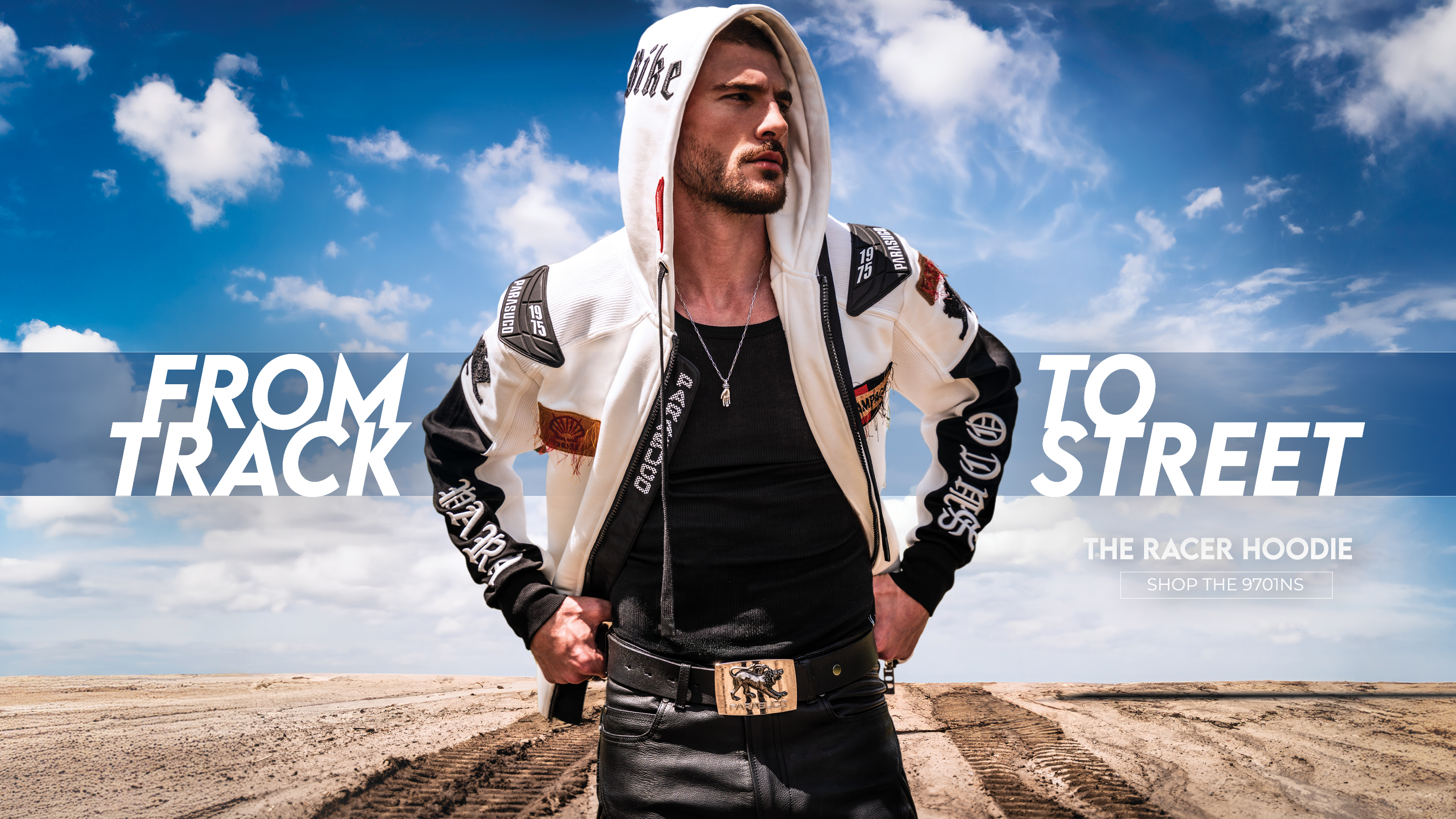 
A muscular man with short hair and tattoos stands in profile, leaning on a rustic wooden fence. He is wearing a black, sleeveless top with a white graphic print, black leather pants, and red motorcycle boots. He holds a red and black helmet in his left hand. The background is a dirt road bordered by trees, with sunlight filtering through the leaves. The image has a dark, vertical overlay on the left side with the text 